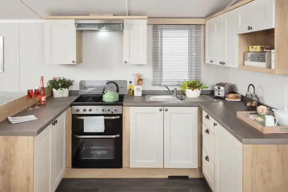 Modern kitchen featuring white cabinets with wooden accents, a stainless steel stove, and a sink. Countertops are dark, and the space includes small plants, dishes, and breakfast items. Natural light filters through a window with blinds.