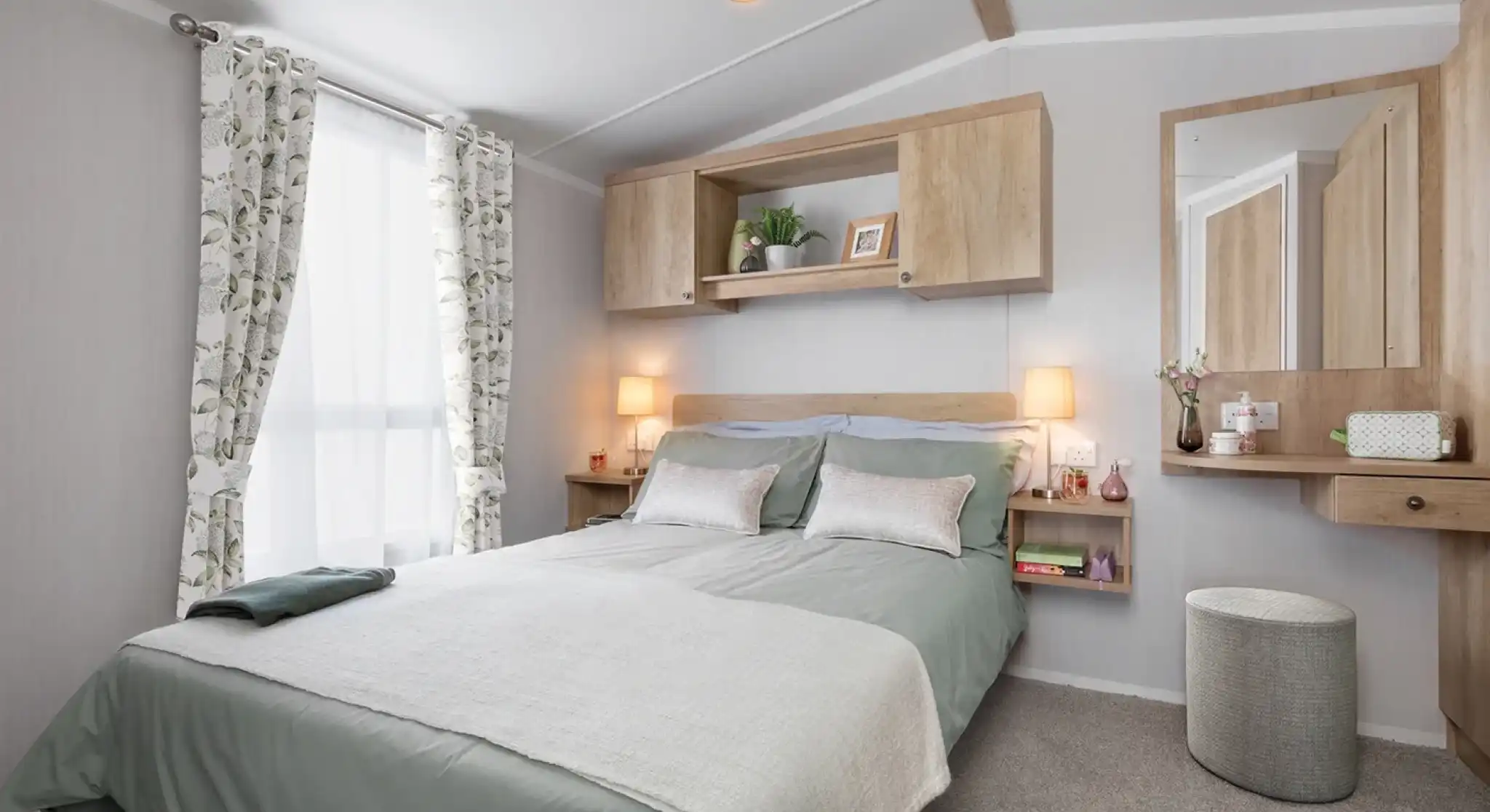 A cozy bedroom featuring a double bed with light green bedding, decorative pillows, and a textured throw. There are wooden shelves above the bed, a stylish mirror on the wall, and soft lighting from bedside lamps. A stool and curtains with a floral pattern complete the inviting atmosphere.