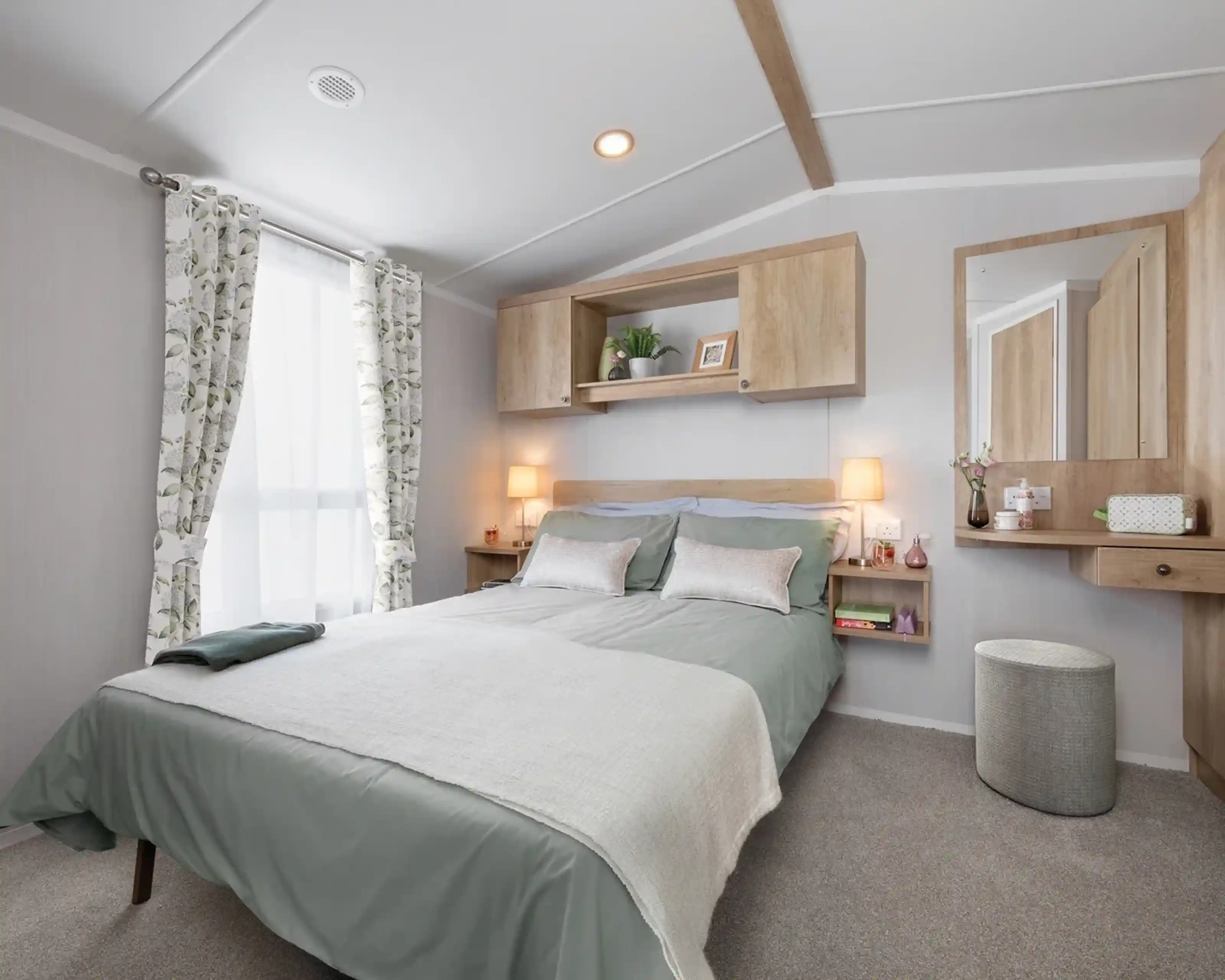 A cozy bedroom featuring a double bed with light green bedding, decorative pillows, and a textured throw. There are wooden shelves above the bed, a stylish mirror on the wall, and soft lighting from bedside lamps. A stool and curtains with a floral pattern complete the inviting atmosphere.