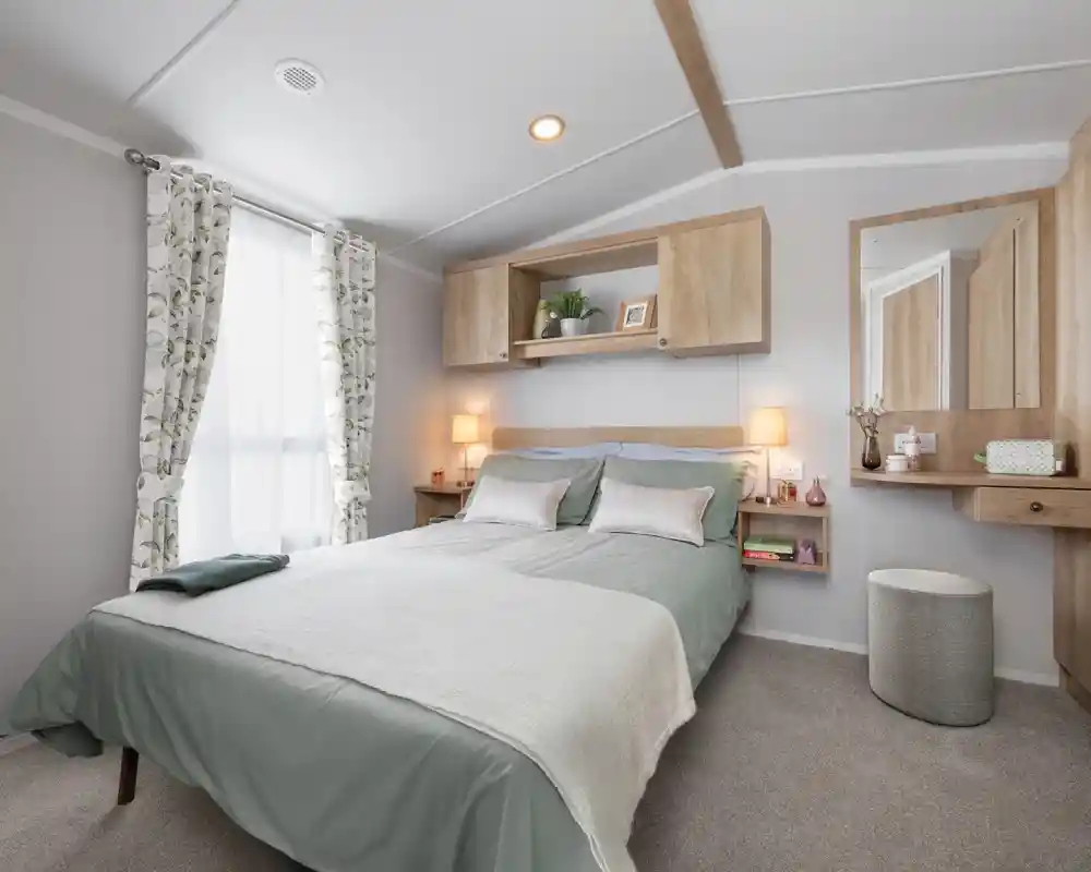 A cozy bedroom featuring a double bed with light green bedding, decorative pillows, and a textured throw. There are wooden shelves above the bed, a stylish mirror on the wall, and soft lighting from bedside lamps. A stool and curtains with a floral pattern complete the inviting atmosphere.