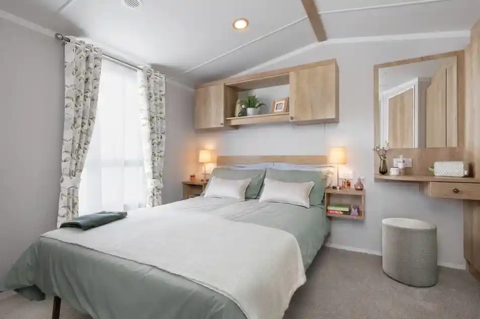 A cozy bedroom featuring a double bed with light green bedding, decorative pillows, and a textured throw. There are wooden shelves above the bed, a stylish mirror on the wall, and soft lighting from bedside lamps. A stool and curtains with a floral pattern complete the inviting atmosphere.