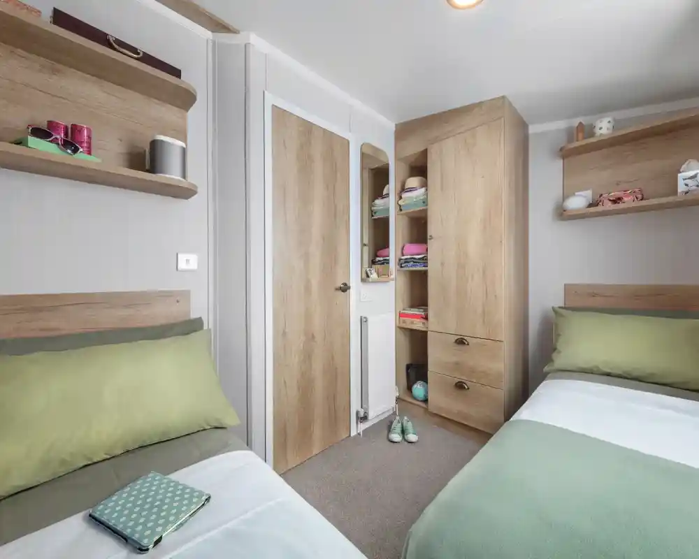 A cozy bedroom with two single beds featuring green and white bedding. There are wooden shelves on the walls holding various items, and a tall wooden cabinet with a closed door. A small door leads to a closet space, and the room has a neutral color scheme with a warm atmosphere.