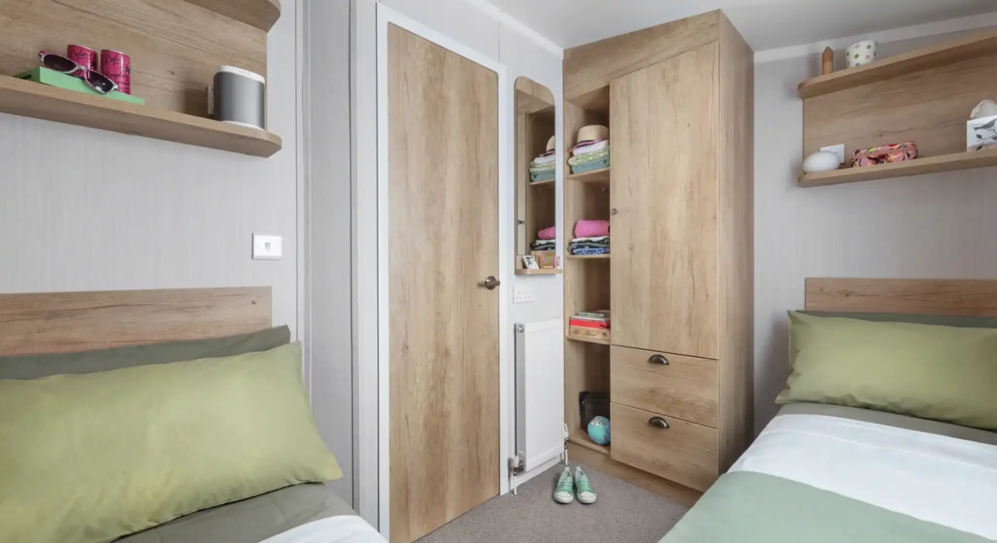 A cozy bedroom with two single beds featuring green and white bedding. There are wooden shelves on the walls holding various items, and a tall wooden cabinet with a closed door. A small door leads to a closet space, and the room has a neutral color scheme with a warm atmosphere.