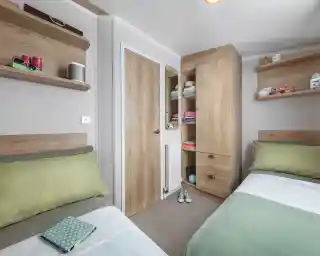 A cozy bedroom with two single beds featuring green and white bedding. There are wooden shelves on the walls holding various items, and a tall wooden cabinet with a closed door. A small door leads to a closet space, and the room has a neutral color scheme with a warm atmosphere.