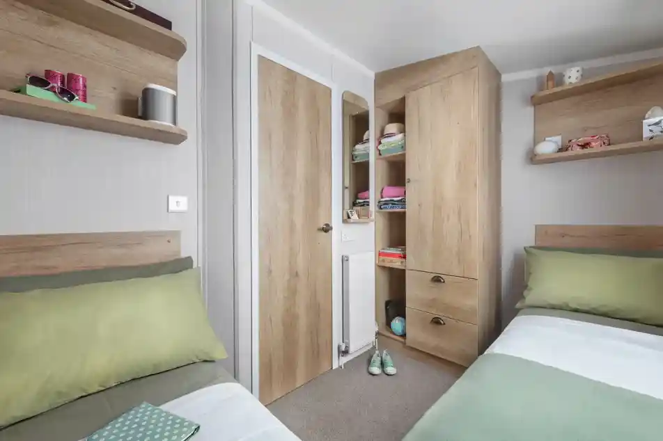 A cozy bedroom with two single beds featuring green and white bedding. There are wooden shelves on the walls holding various items, and a tall wooden cabinet with a closed door. A small door leads to a closet space, and the room has a neutral color scheme with a warm atmosphere.