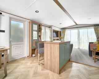 View of the ABI Ingleton holiday home's entryway and open-plan living area, featuring a built-in breakfast bar with sage green paneling and wood accents, paired with two upholstered bar stools. The area leads into a bright lounge with large sliding glass doors and floral curtains. Light wood herringbone flooring contrasts with soft grey carpet in the living space. A hallway bench with storage, mirror, and coat hooks adds functionality near the entrance.