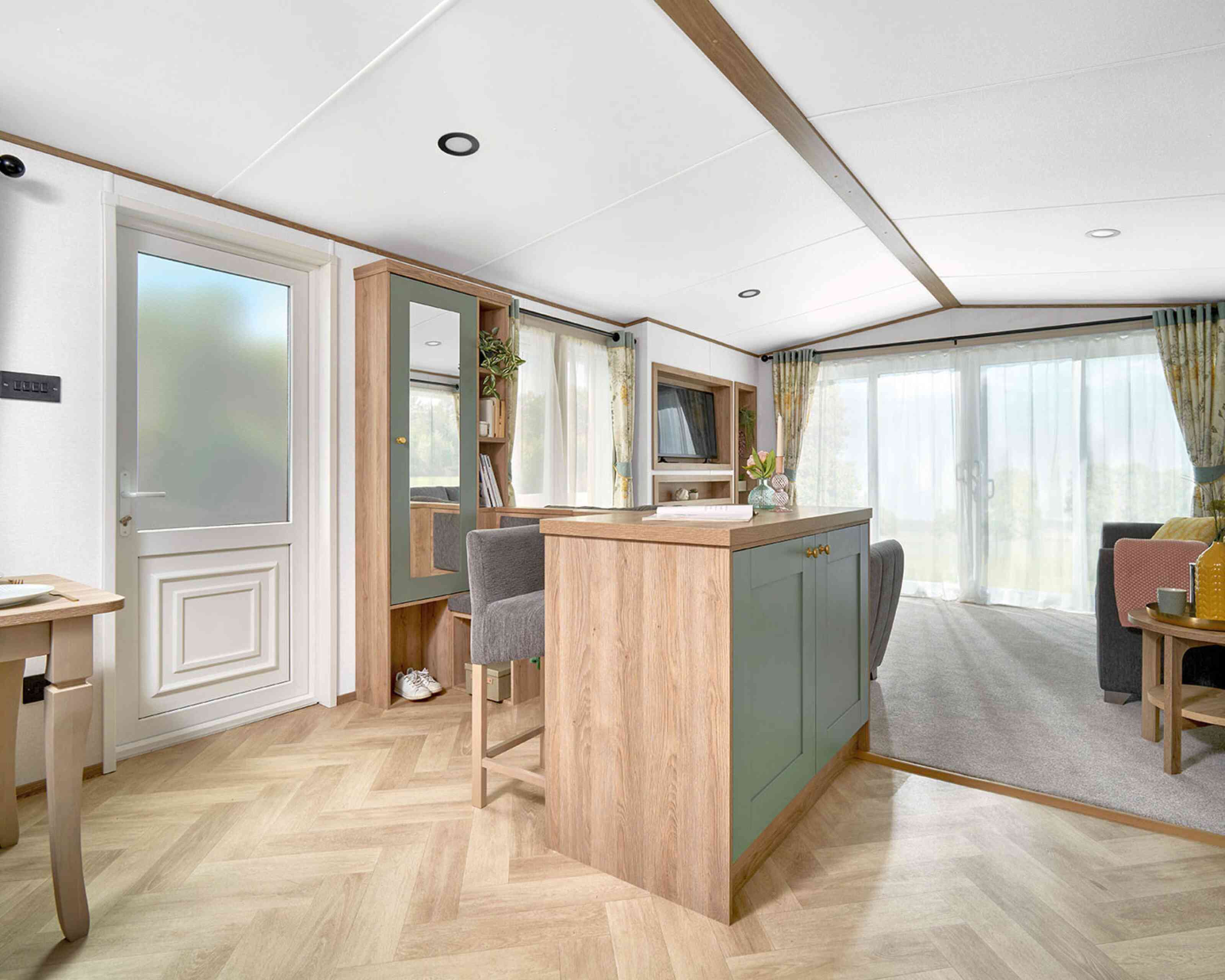 View of the ABI Ingleton holiday home's entryway and open-plan living area, featuring a built-in breakfast bar with sage green paneling and wood accents, paired with two upholstered bar stools. The area leads into a bright lounge with large sliding glass doors and floral curtains. Light wood herringbone flooring contrasts with soft grey carpet in the living space. A hallway bench with storage, mirror, and coat hooks adds functionality near the entrance.
