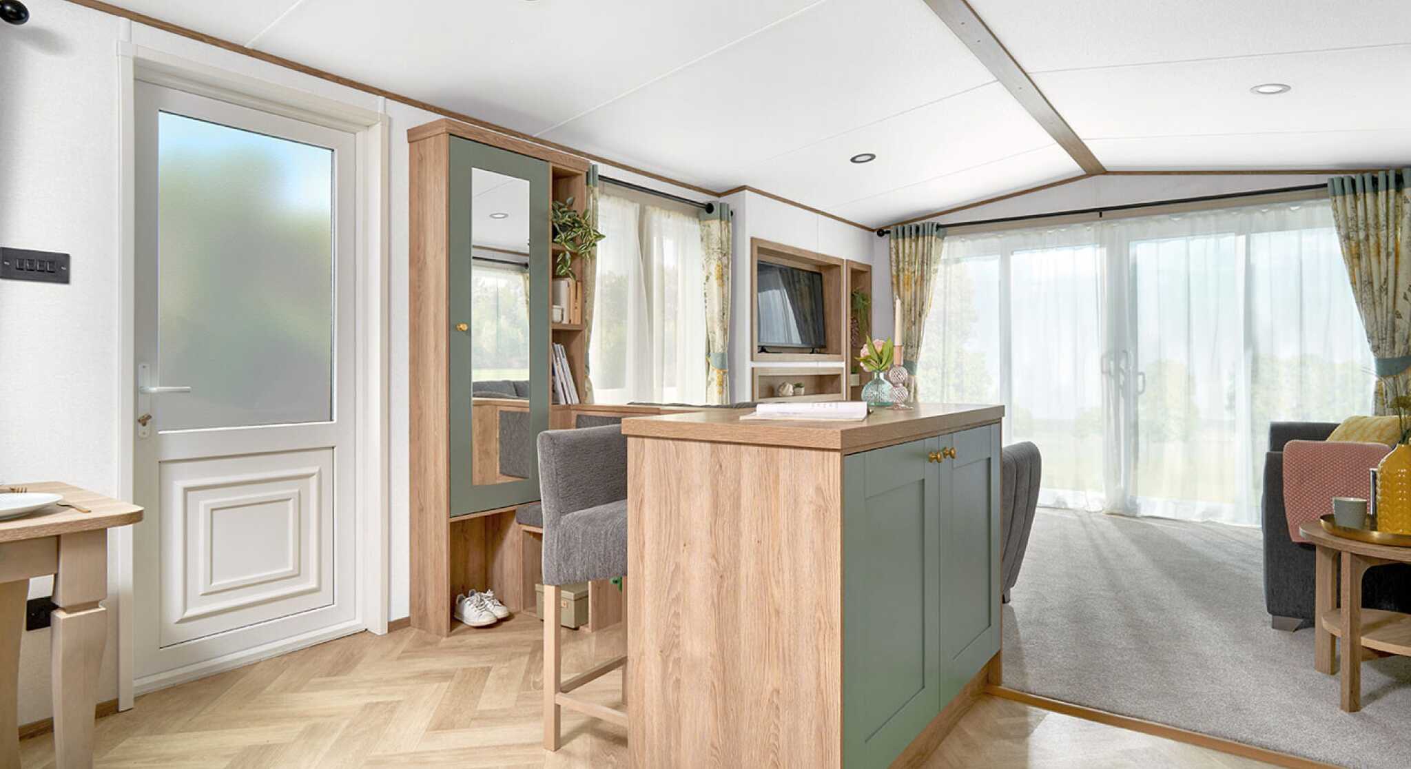View of the ABI Ingleton holiday home's entryway and open-plan living area, featuring a built-in breakfast bar with sage green paneling and wood accents, paired with two upholstered bar stools. The area leads into a bright lounge with large sliding glass doors and floral curtains. Light wood herringbone flooring contrasts with soft grey carpet in the living space. A hallway bench with storage, mirror, and coat hooks adds functionality near the entrance.