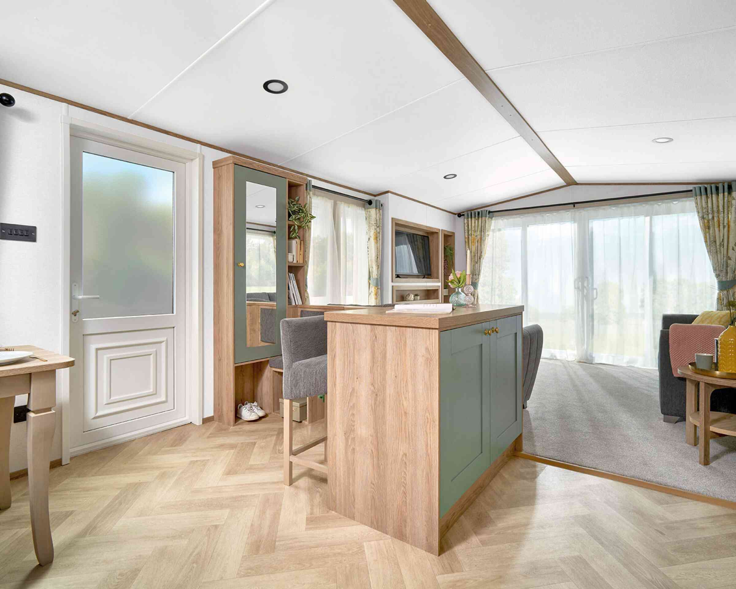 View of the ABI Ingleton holiday home's entryway and open-plan living area, featuring a built-in breakfast bar with sage green paneling and wood accents, paired with two upholstered bar stools. The area leads into a bright lounge with large sliding glass doors and floral curtains. Light wood herringbone flooring contrasts with soft grey carpet in the living space. A hallway bench with storage, mirror, and coat hooks adds functionality near the entrance.