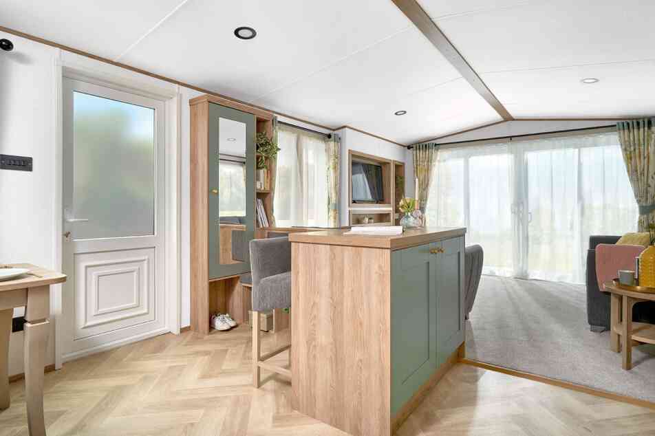 View of the ABI Ingleton holiday home's entryway and open-plan living area, featuring a built-in breakfast bar with sage green paneling and wood accents, paired with two upholstered bar stools. The area leads into a bright lounge with large sliding glass doors and floral curtains. Light wood herringbone flooring contrasts with soft grey carpet in the living space. A hallway bench with storage, mirror, and coat hooks adds functionality near the entrance.