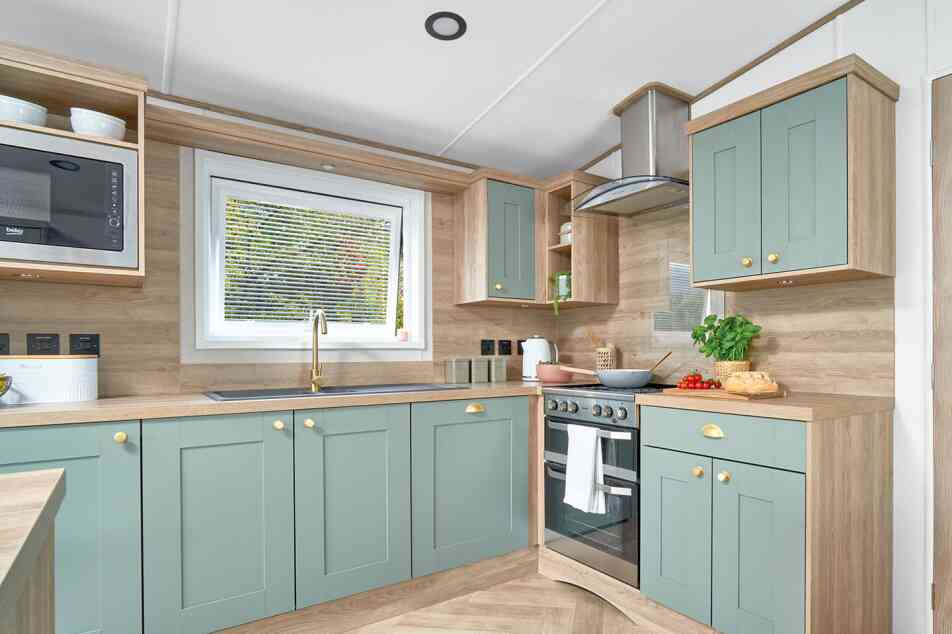 Contemporary L-shaped kitchen inside the ABI Ingleton holiday home, featuring soft sage green shaker-style cabinets with gold handles, light wood countertops, and matching wood panel walls. A window above the sink allows natural light in, while a built-in oven with a glass hob and stainless steel extractor hood completes the modern look. Open shelving and built-in microwave add convenience and storage, with fresh produce and kitchen items neatly displayed on the counters.