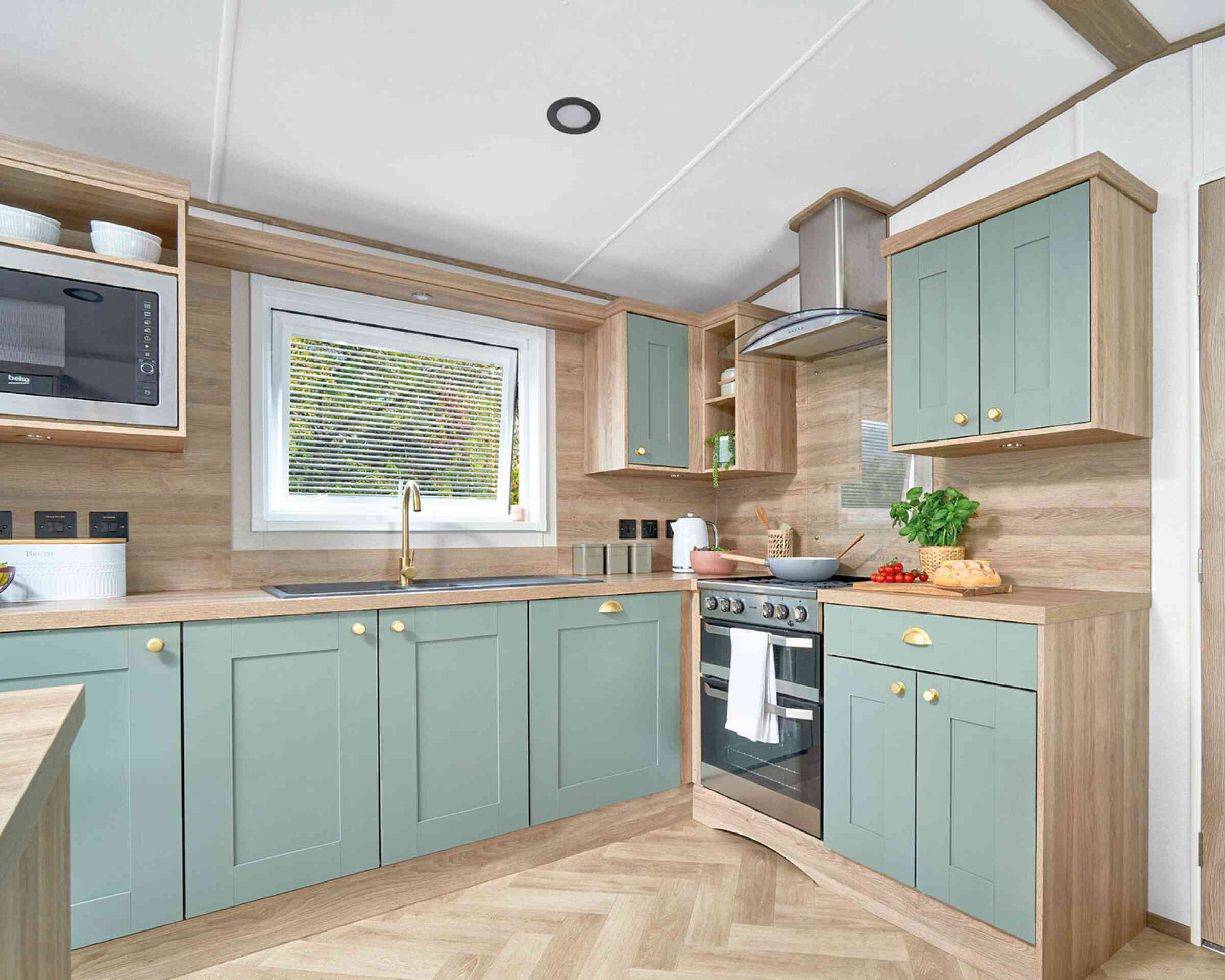 Contemporary L-shaped kitchen inside the ABI Ingleton holiday home, featuring soft sage green shaker-style cabinets with gold handles, light wood countertops, and matching wood panel walls. A window above the sink allows natural light in, while a built-in oven with a glass hob and stainless steel extractor hood completes the modern look. Open shelving and built-in microwave add convenience and storage, with fresh produce and kitchen items neatly displayed on the counters.