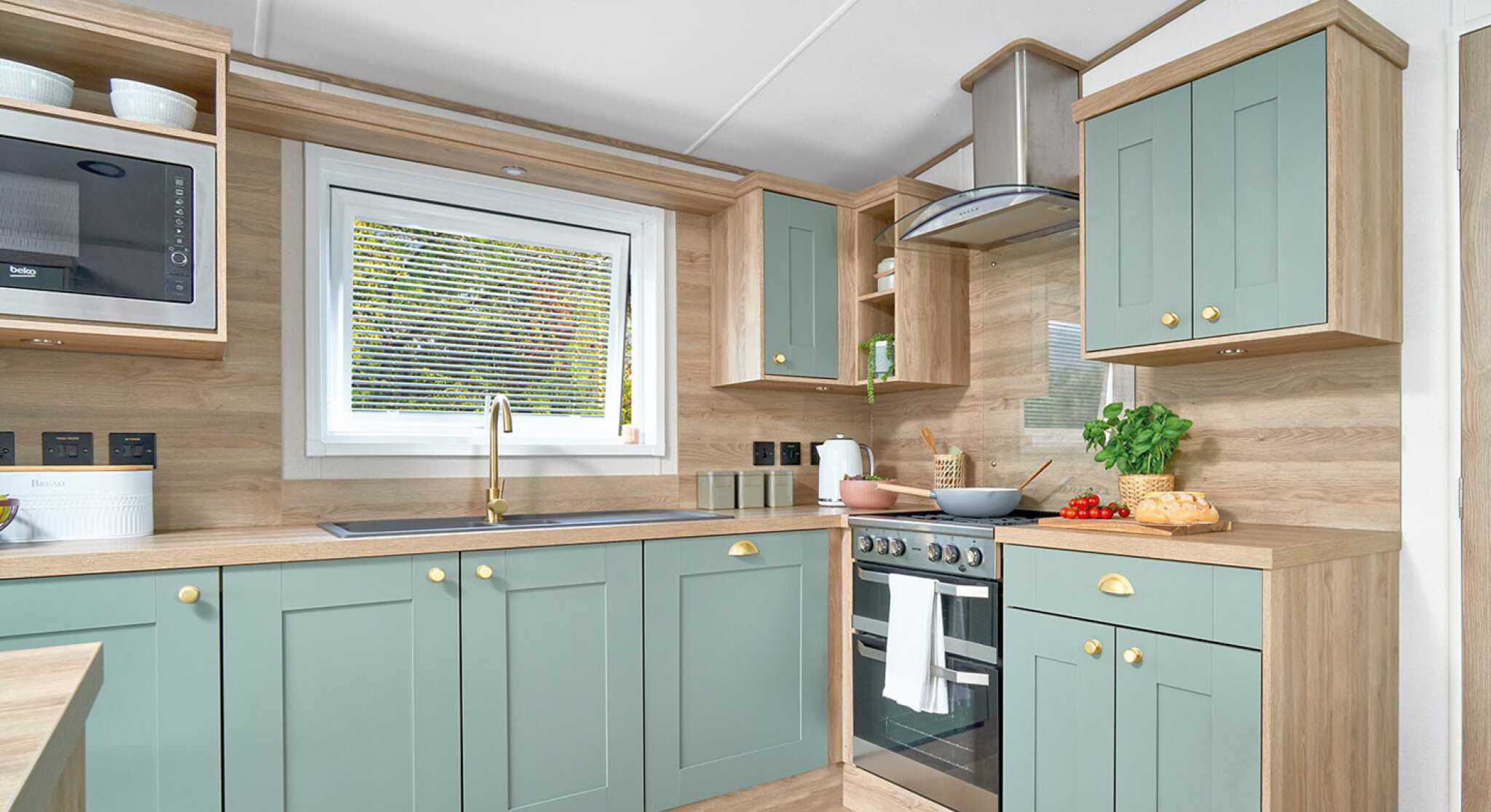 Contemporary L-shaped kitchen inside the ABI Ingleton holiday home, featuring soft sage green shaker-style cabinets with gold handles, light wood countertops, and matching wood panel walls. A window above the sink allows natural light in, while a built-in oven with a glass hob and stainless steel extractor hood completes the modern look. Open shelving and built-in microwave add convenience and storage, with fresh produce and kitchen items neatly displayed on the counters.