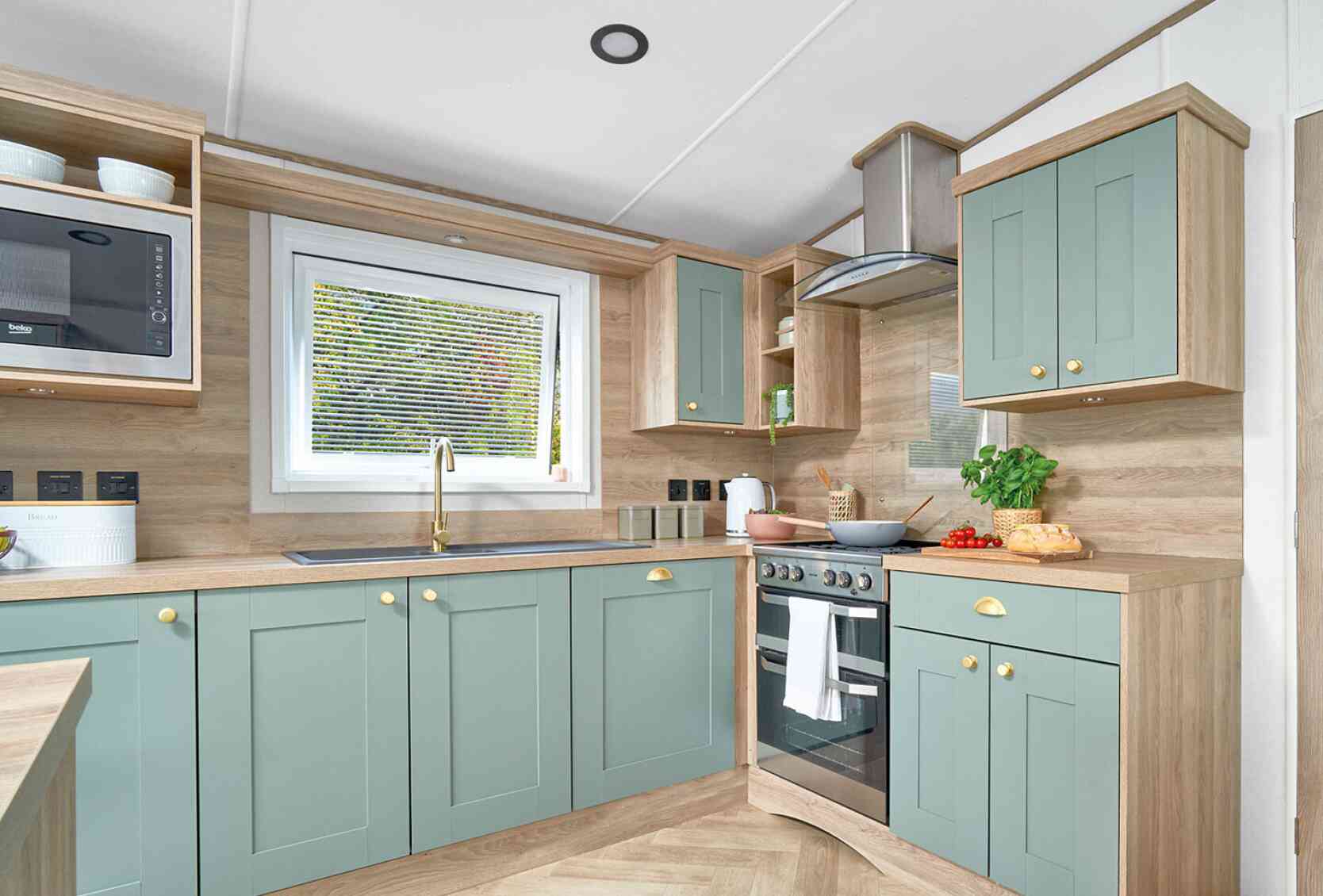 Contemporary L-shaped kitchen inside the ABI Ingleton holiday home, featuring soft sage green shaker-style cabinets with gold handles, light wood countertops, and matching wood panel walls. A window above the sink allows natural light in, while a built-in oven with a glass hob and stainless steel extractor hood completes the modern look. Open shelving and built-in microwave add convenience and storage, with fresh produce and kitchen items neatly displayed on the counters.