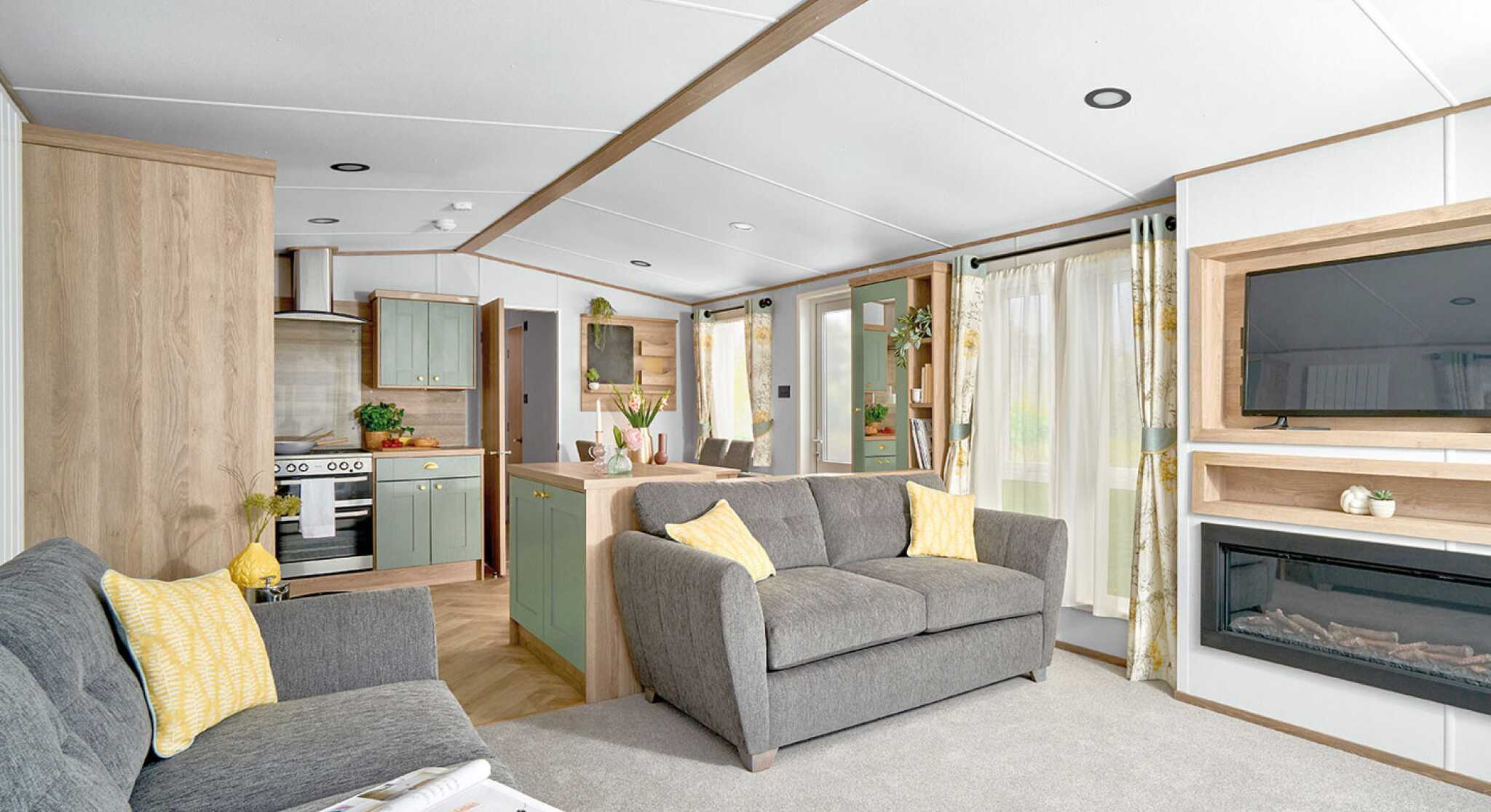 Modern open-plan living area inside the ABI Ingleton holiday home, featuring two grey sofas with yellow cushions on a light grey carpet. A wall-mounted TV sits above a built-in electric fireplace with shelving. The space flows into a stylish kitchen with sage green cabinets, wood accents, and a double oven. Large windows with floral curtains allow natural light to brighten the room, creating a cozy and inviting atmosphere.