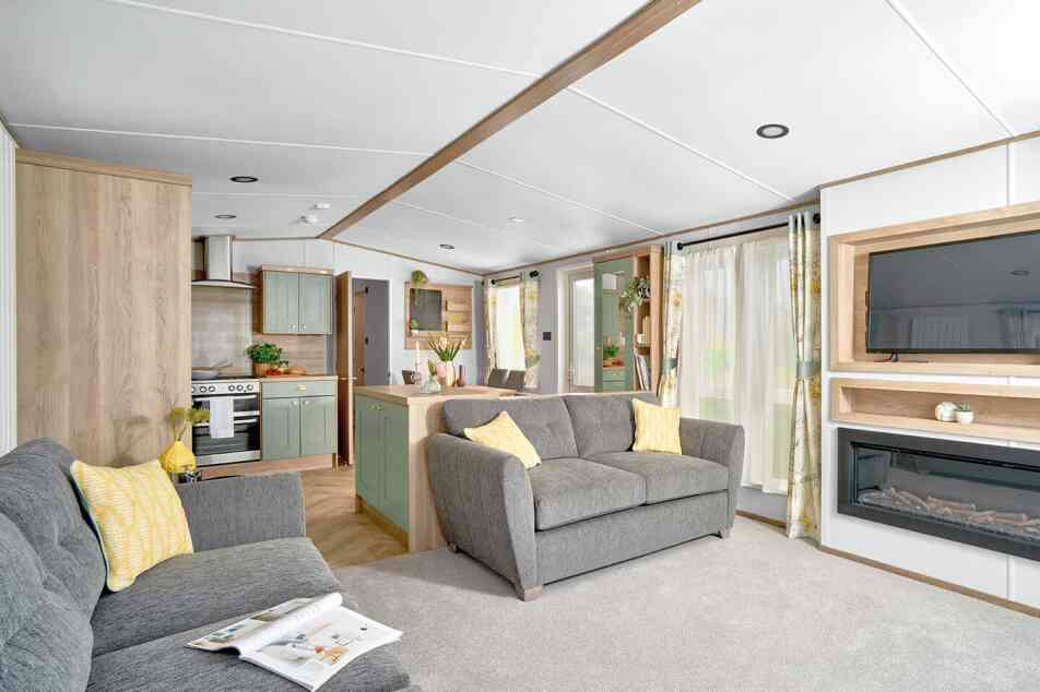 Modern open-plan living area inside the ABI Ingleton holiday home, featuring two grey sofas with yellow cushions on a light grey carpet. A wall-mounted TV sits above a built-in electric fireplace with shelving. The space flows into a stylish kitchen with sage green cabinets, wood accents, and a double oven. Large windows with floral curtains allow natural light to brighten the room, creating a cozy and inviting atmosphere.