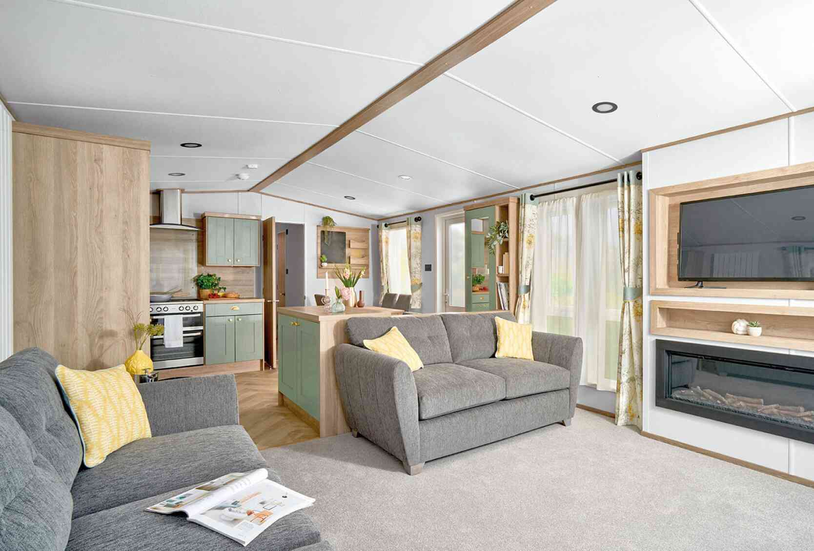 Modern open-plan living area inside the ABI Ingleton holiday home, featuring two grey sofas with yellow cushions on a light grey carpet. A wall-mounted TV sits above a built-in electric fireplace with shelving. The space flows into a stylish kitchen with sage green cabinets, wood accents, and a double oven. Large windows with floral curtains allow natural light to brighten the room, creating a cozy and inviting atmosphere.