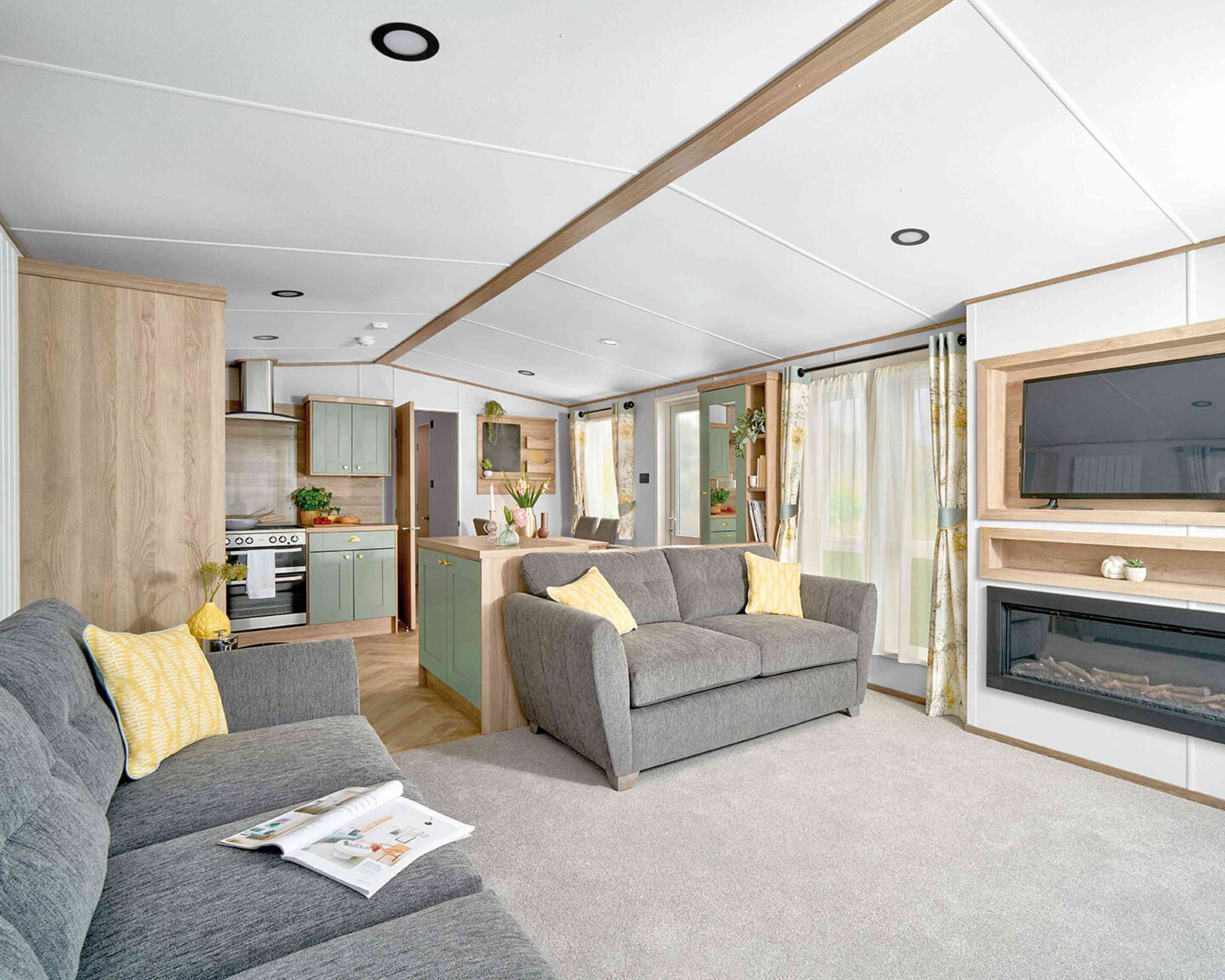 Modern open-plan living area inside the ABI Ingleton holiday home, featuring two grey sofas with yellow cushions on a light grey carpet. A wall-mounted TV sits above a built-in electric fireplace with shelving. The space flows into a stylish kitchen with sage green cabinets, wood accents, and a double oven. Large windows with floral curtains allow natural light to brighten the room, creating a cozy and inviting atmosphere.