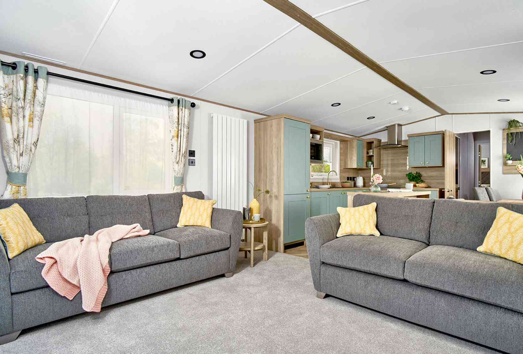 Bright and spacious lounge area of the ABI Ingleton holiday home featuring two large grey sofas with yellow cushions, a soft grey carpet, and a side table with decorative items. The open-plan layout leads into a modern kitchen with mint green cabinets, wood finishes, and integrated appliances. Natural light fills the space through large windows with patterned curtains.