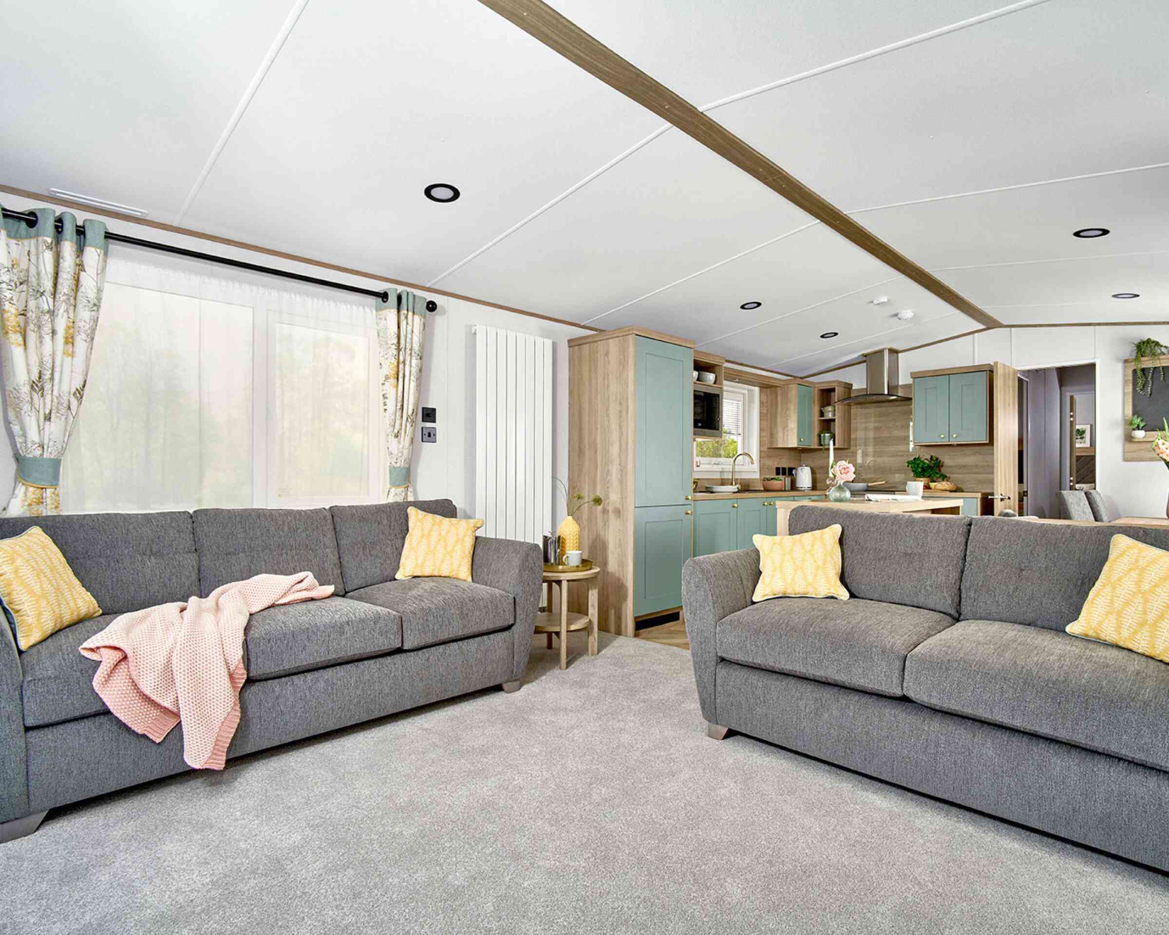 Bright and spacious lounge area of the ABI Ingleton holiday home featuring two large grey sofas with yellow cushions, a soft grey carpet, and a side table with decorative items. The open-plan layout leads into a modern kitchen with mint green cabinets, wood finishes, and integrated appliances. Natural light fills the space through large windows with patterned curtains.