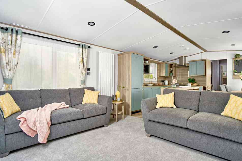 Bright and spacious lounge area of the ABI Ingleton holiday home featuring two large grey sofas with yellow cushions, a soft grey carpet, and a side table with decorative items. The open-plan layout leads into a modern kitchen with mint green cabinets, wood finishes, and integrated appliances. Natural light fills the space through large windows with patterned curtains.