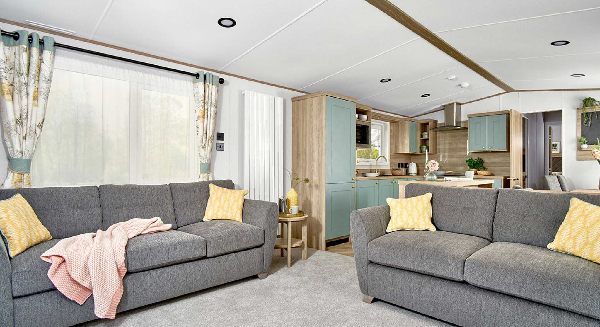 Bright and spacious lounge area of the ABI Ingleton holiday home featuring two large grey sofas with yellow cushions, a soft grey carpet, and a side table with decorative items. The open-plan layout leads into a modern kitchen with mint green cabinets, wood finishes, and integrated appliances. Natural light fills the space through large windows with patterned curtains.