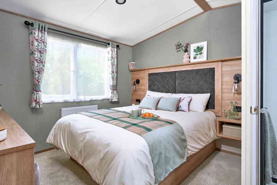 Master bedroom inside the ABI Ingleton holiday home featuring a double bed with a large upholstered headboard set in a wood surround. The bed is neatly made with white bedding, floral accent pillows, and a soft green and plaid runner. Floating bedside tables with lamps and glassware add convenience. A window with floral curtains lets in natural light, and a dresser with a cushioned stool sits opposite the bed. Soft carpeting and decorative touches create a calm, cosy atmosphere.