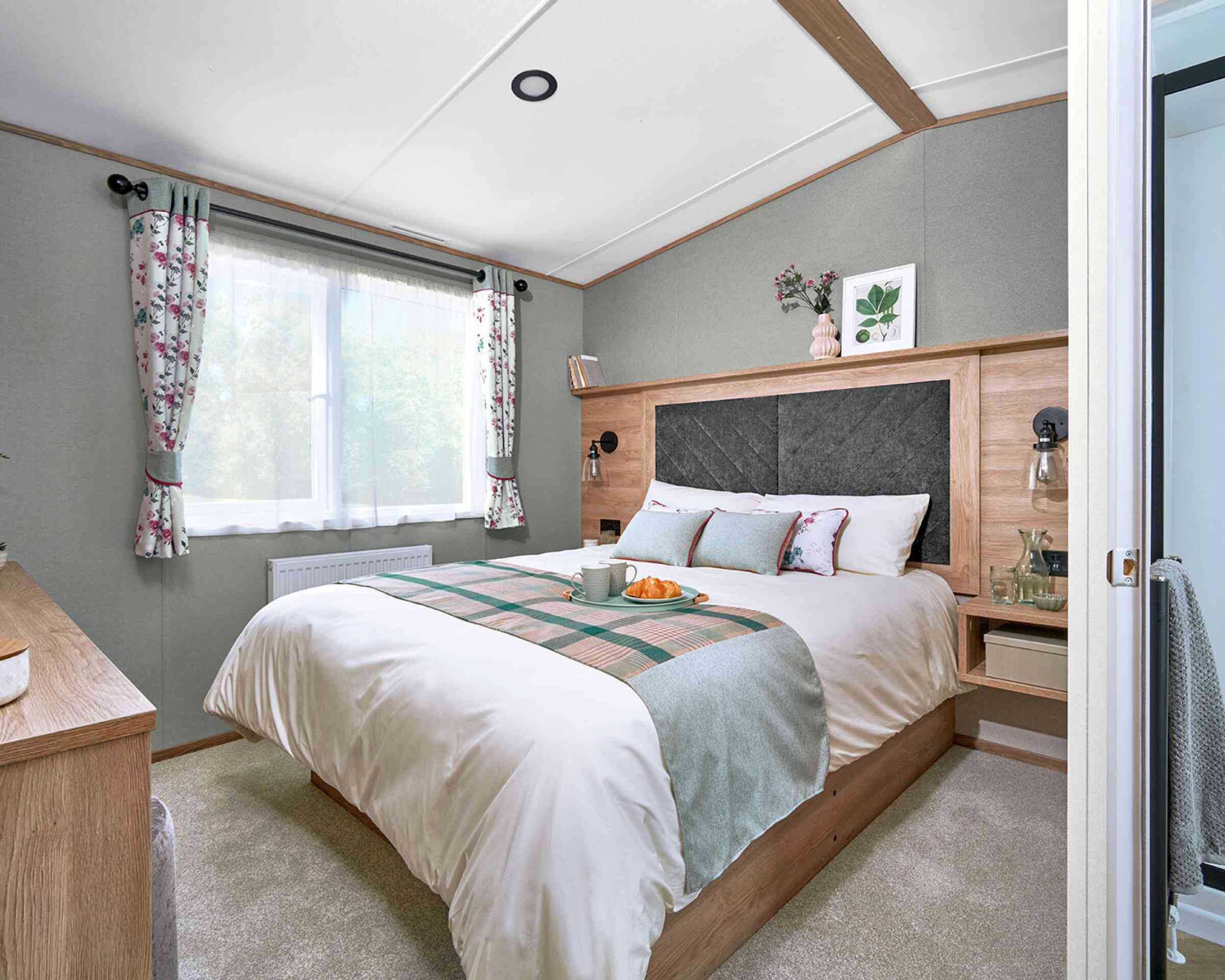 Master bedroom inside the ABI Ingleton holiday home featuring a double bed with a large upholstered headboard set in a wood surround. The bed is neatly made with white bedding, floral accent pillows, and a soft green and plaid runner. Floating bedside tables with lamps and glassware add convenience. A window with floral curtains lets in natural light, and a dresser with a cushioned stool sits opposite the bed. Soft carpeting and decorative touches create a calm, cosy atmosphere.