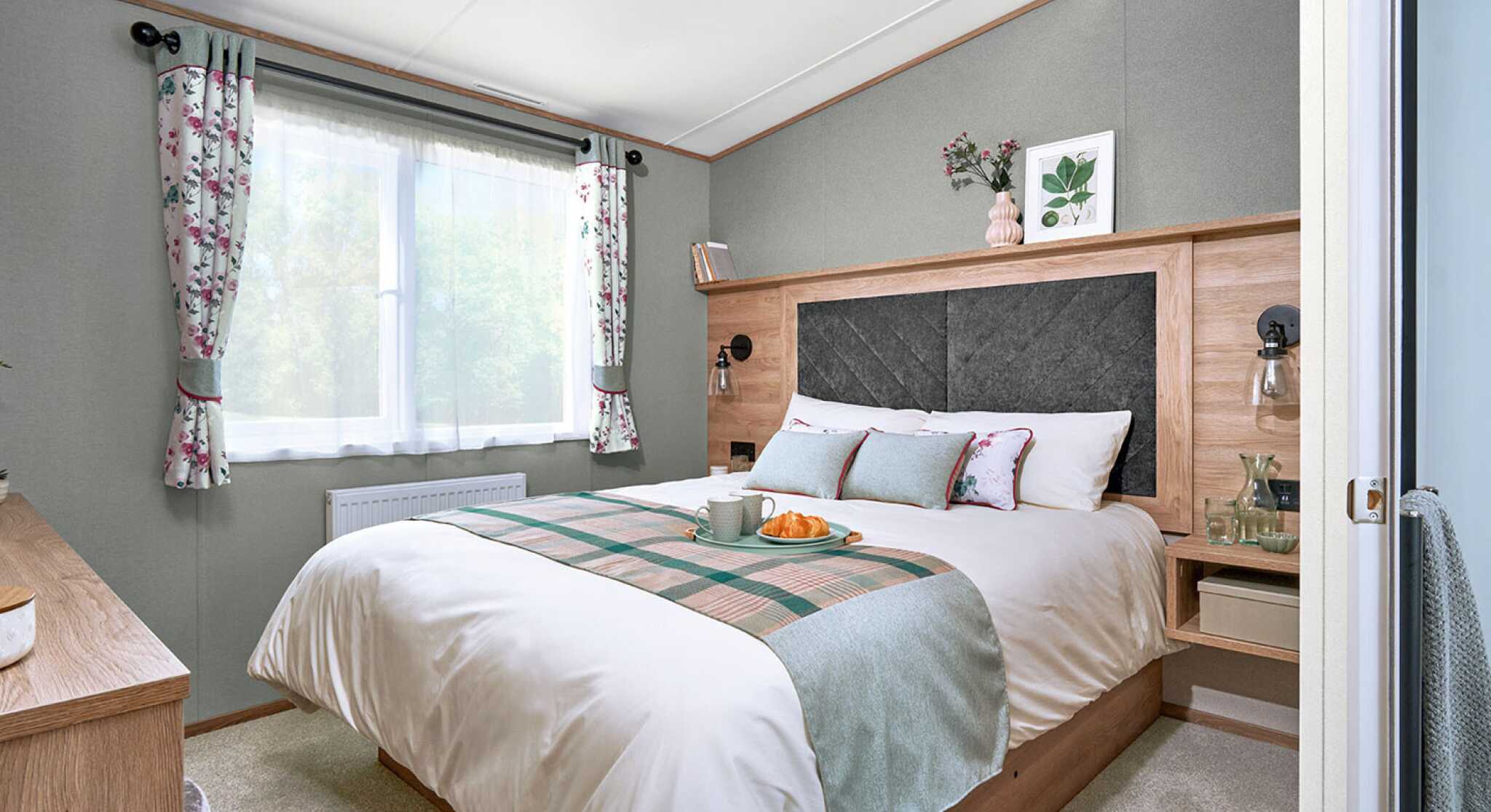 Master bedroom inside the ABI Ingleton holiday home featuring a double bed with a large upholstered headboard set in a wood surround. The bed is neatly made with white bedding, floral accent pillows, and a soft green and plaid runner. Floating bedside tables with lamps and glassware add convenience. A window with floral curtains lets in natural light, and a dresser with a cushioned stool sits opposite the bed. Soft carpeting and decorative touches create a calm, cosy atmosphere.