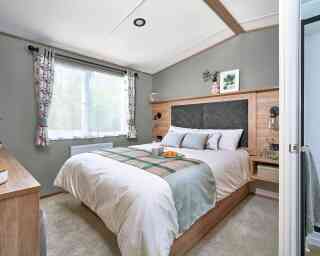 Master bedroom inside the ABI Ingleton holiday home featuring a double bed with a large upholstered headboard set in a wood surround. The bed is neatly made with white bedding, floral accent pillows, and a soft green and plaid runner. Floating bedside tables with lamps and glassware add convenience. A window with floral curtains lets in natural light, and a dresser with a cushioned stool sits opposite the bed. Soft carpeting and decorative touches create a calm, cosy atmosphere.