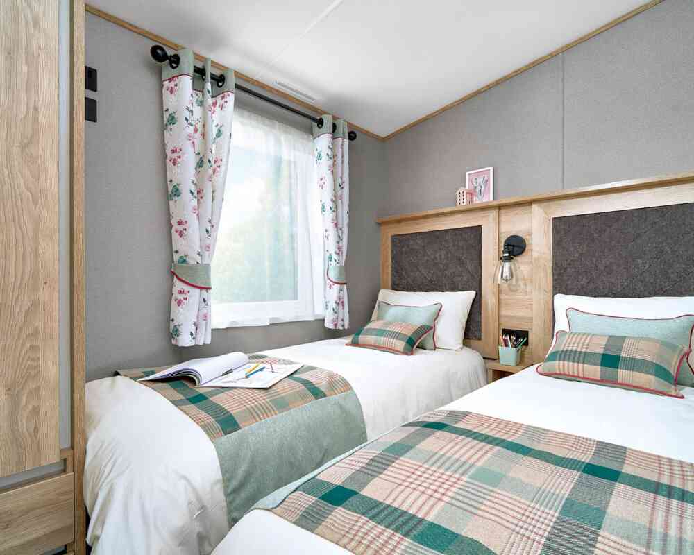 Twin bedroom inside the ABI Ingleton holiday home, featuring two single beds with matching green and plaid runners, decorative cushions, and upholstered headboards framed in wood. A shared nightstand with a lamp and storage sits between the beds. Light enters through a window dressed with floral curtains, complementing the soft grey walls and cozy atmosphere. A wardrobe is partially visible on the left, offering practical storage.