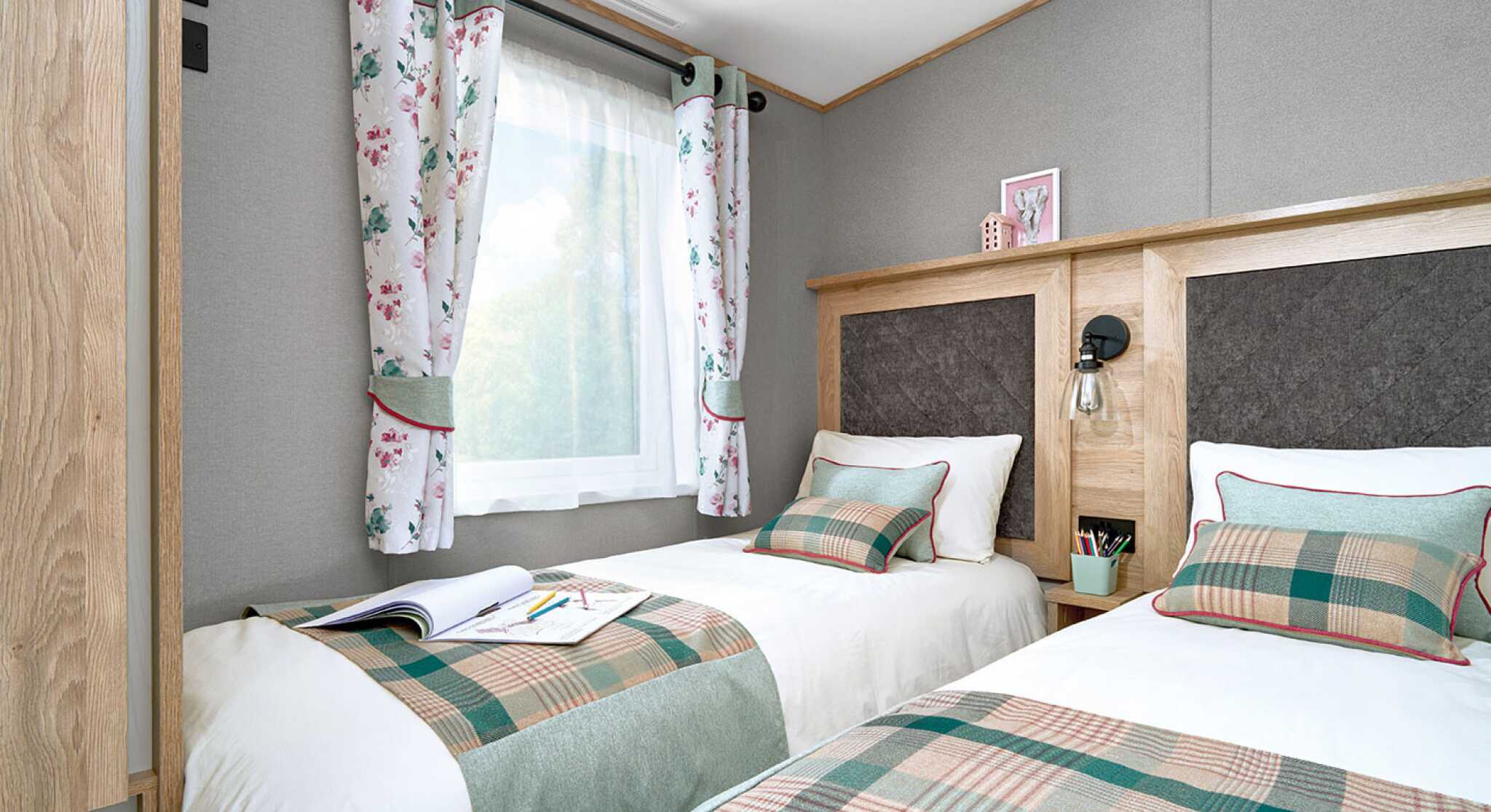 Twin bedroom inside the ABI Ingleton holiday home, featuring two single beds with matching green and plaid runners, decorative cushions, and upholstered headboards framed in wood. A shared nightstand with a lamp and storage sits between the beds. Light enters through a window dressed with floral curtains, complementing the soft grey walls and cozy atmosphere. A wardrobe is partially visible on the left, offering practical storage.