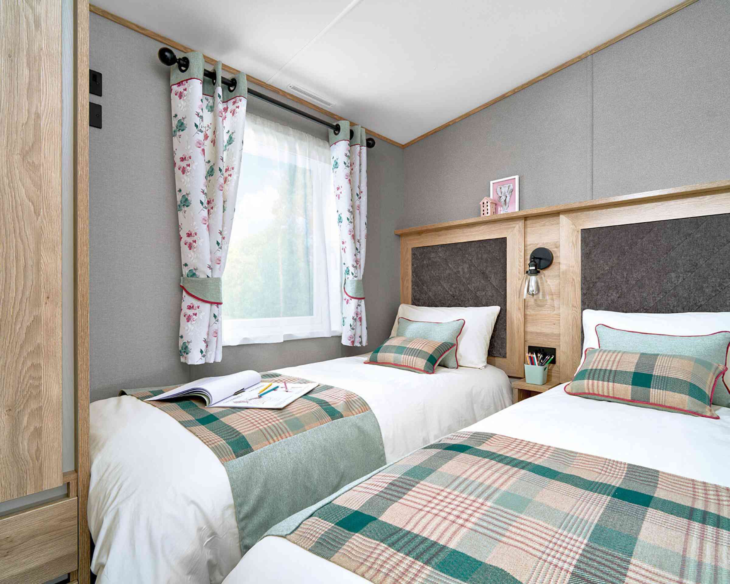 Twin bedroom inside the ABI Ingleton holiday home, featuring two single beds with matching green and plaid runners, decorative cushions, and upholstered headboards framed in wood. A shared nightstand with a lamp and storage sits between the beds. Light enters through a window dressed with floral curtains, complementing the soft grey walls and cozy atmosphere. A wardrobe is partially visible on the left, offering practical storage.