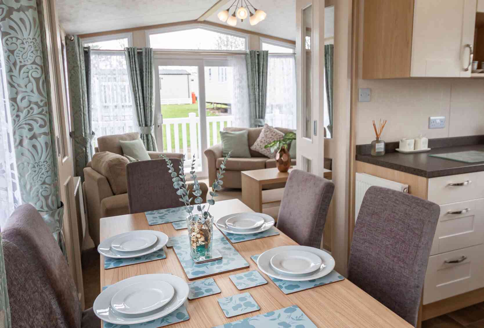 Bright dining and living area in a mobile home, featuring a set table with blue floral placemats, cushioned chairs, sofa, and large windows with curtains.