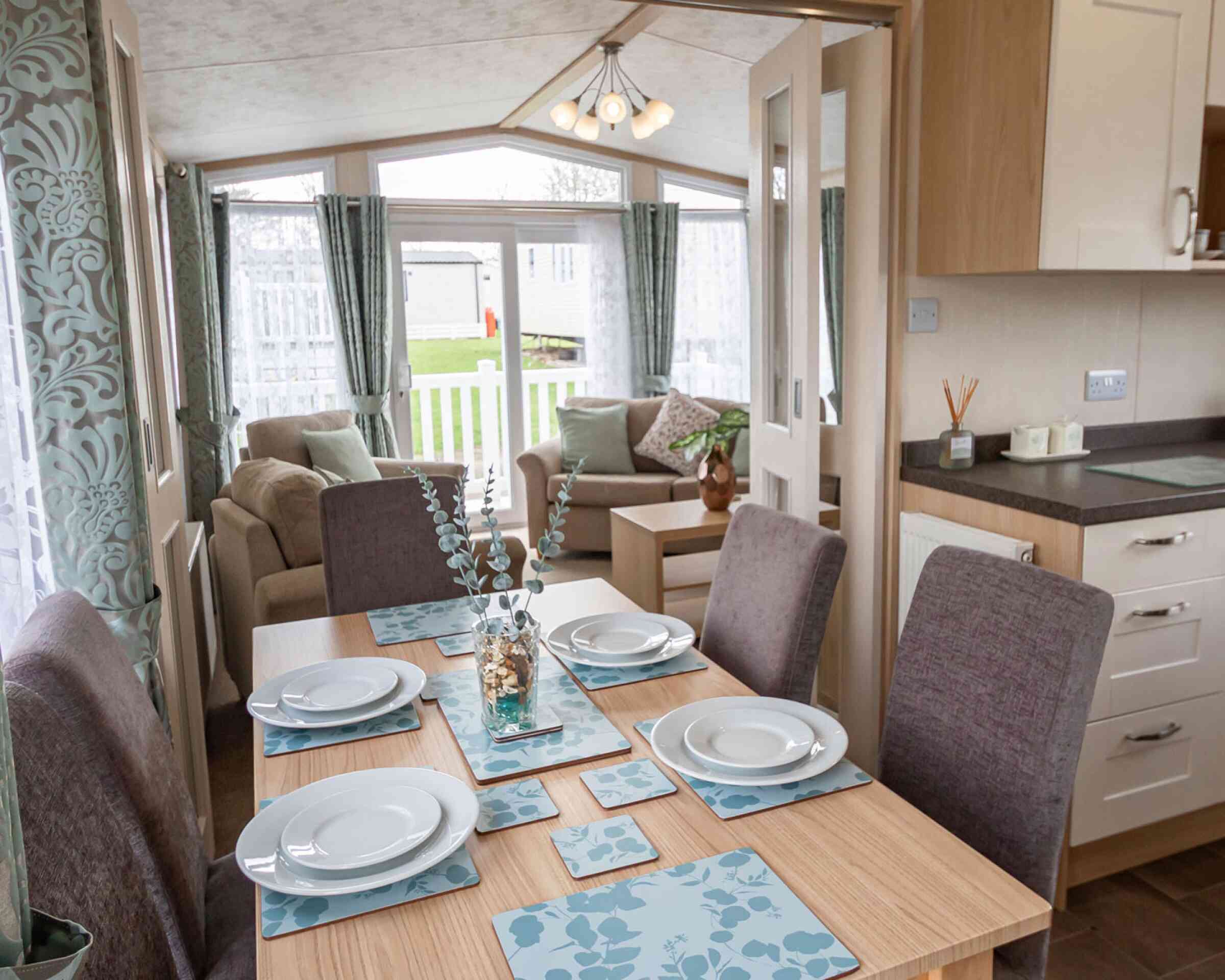Bright dining and living area in a mobile home, featuring a set table with blue floral placemats, cushioned chairs, sofa, and large windows with curtains.