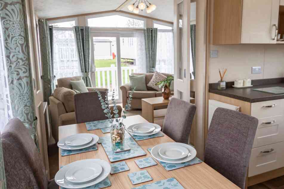 Bright dining and living area in a mobile home, featuring a set table with blue floral placemats, cushioned chairs, sofa, and large windows with curtains.