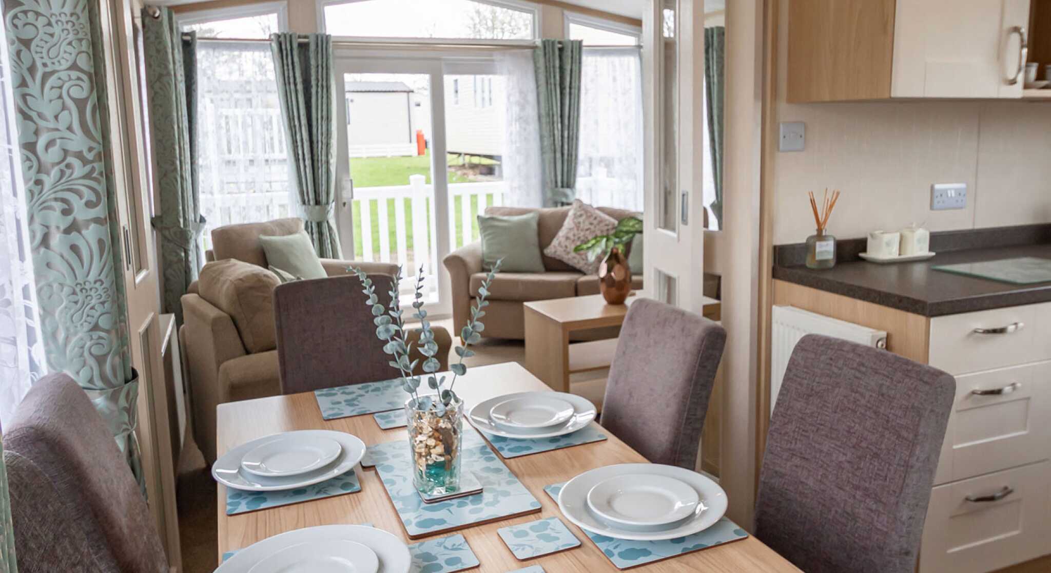 Bright dining and living area in a mobile home, featuring a set table with blue floral placemats, cushioned chairs, sofa, and large windows with curtains.