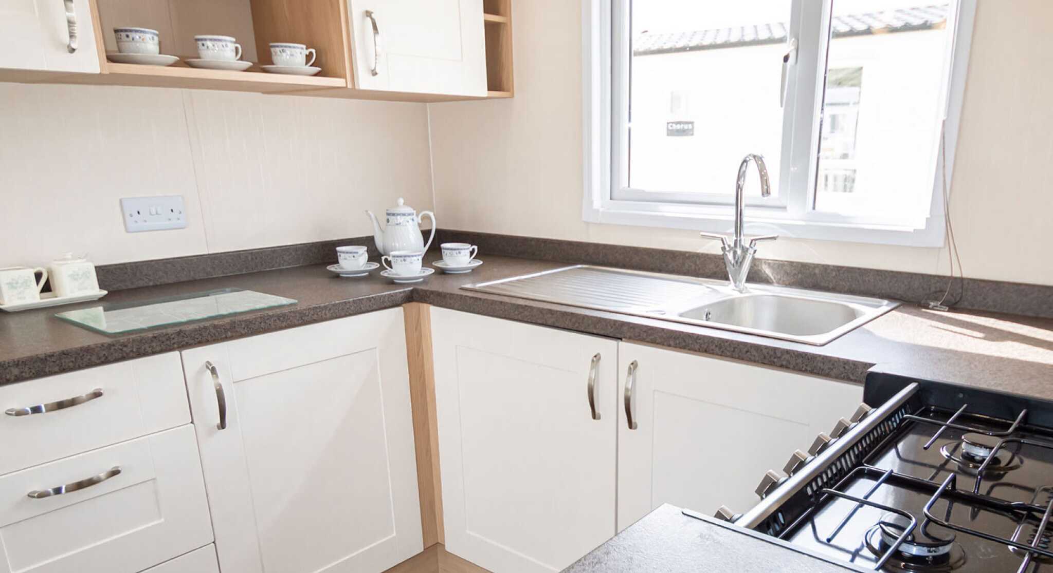 Bright kitchen with white cabinetry and dark countertops. A window over the sink lets in natural light. Teacups and a teapot sit neatly on the counter.