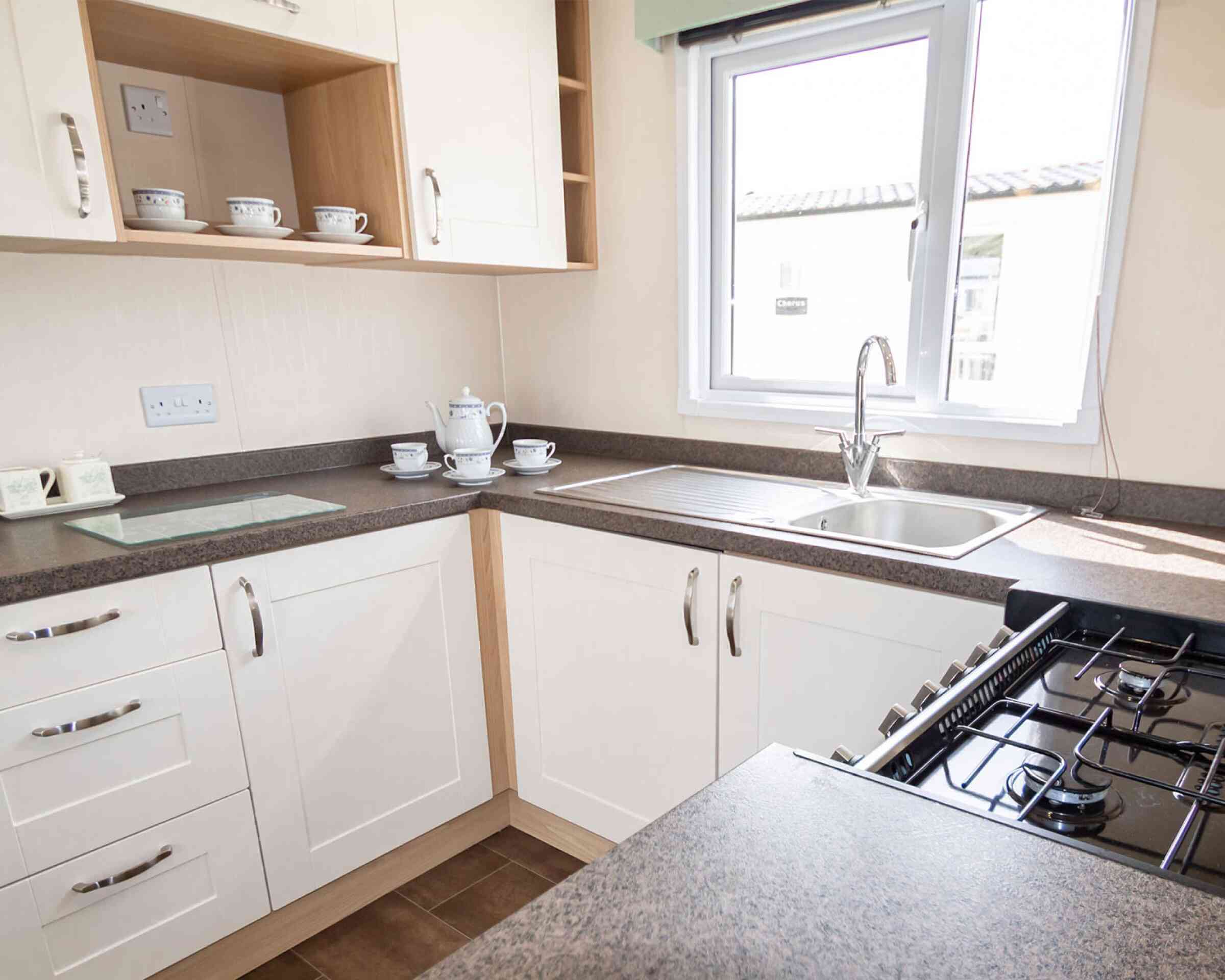Bright kitchen with white cabinetry and dark countertops. A window over the sink lets in natural light. Teacups and a teapot sit neatly on the counter.