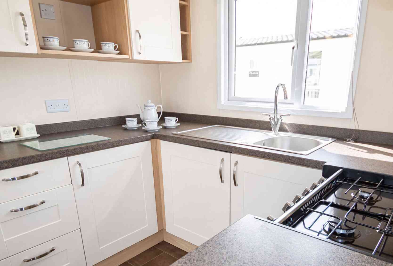 Bright kitchen with white cabinetry and dark countertops. A window over the sink lets in natural light. Teacups and a teapot sit neatly on the counter.