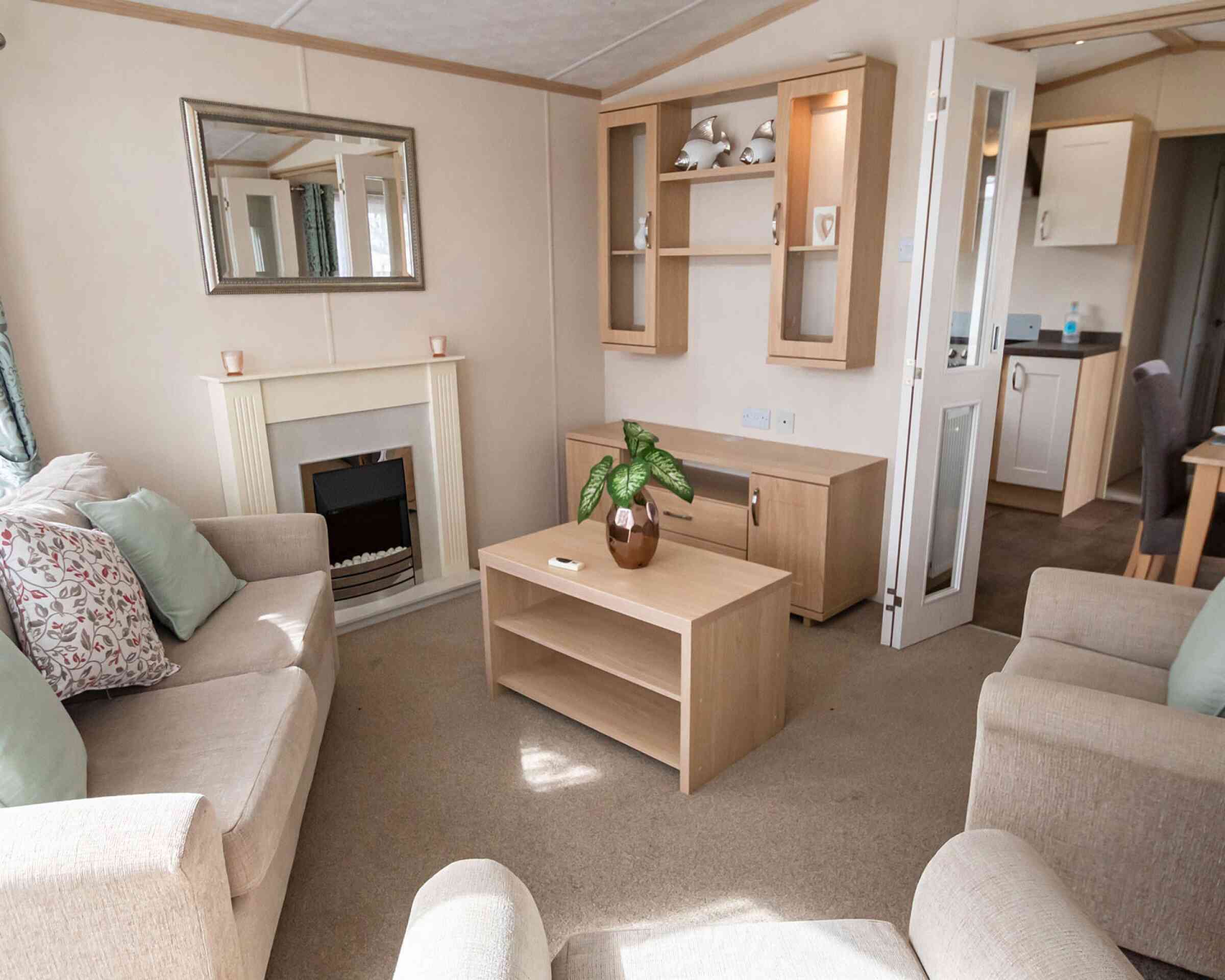 Cosy living room with beige sofas and pastel cushions around a wooden coffee table. A fireplace, mirror, and shelves add a warm, inviting feel.