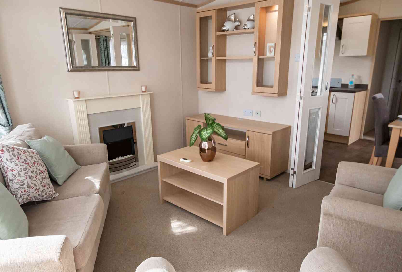 Cosy living room with beige sofas and pastel cushions around a wooden coffee table. A fireplace, mirror, and shelves add a warm, inviting feel.