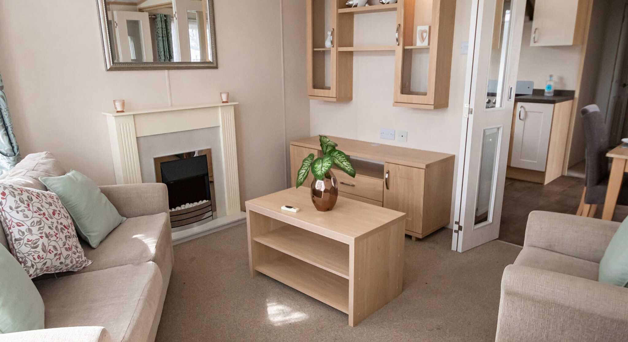 Cosy living room with beige sofas and pastel cushions around a wooden coffee table. A fireplace, mirror, and shelves add a warm, inviting feel.