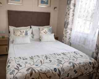 Cosy bedroom with a large bed featuring patterned floral bedding and pillows. Soft, natural light filters through white lace curtains, creating a calm ambiance.