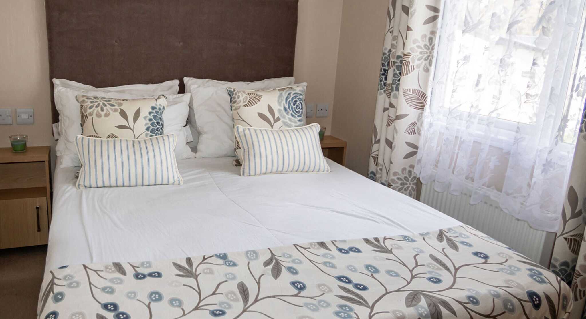 Cosy bedroom with a large bed featuring patterned floral bedding and pillows. Soft, natural light filters through white lace curtains, creating a calm ambiance.