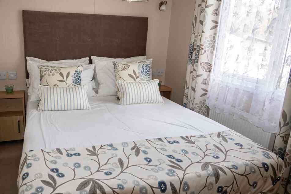 Cosy bedroom with a large bed featuring patterned floral bedding and pillows. Soft, natural light filters through white lace curtains, creating a calm ambiance.