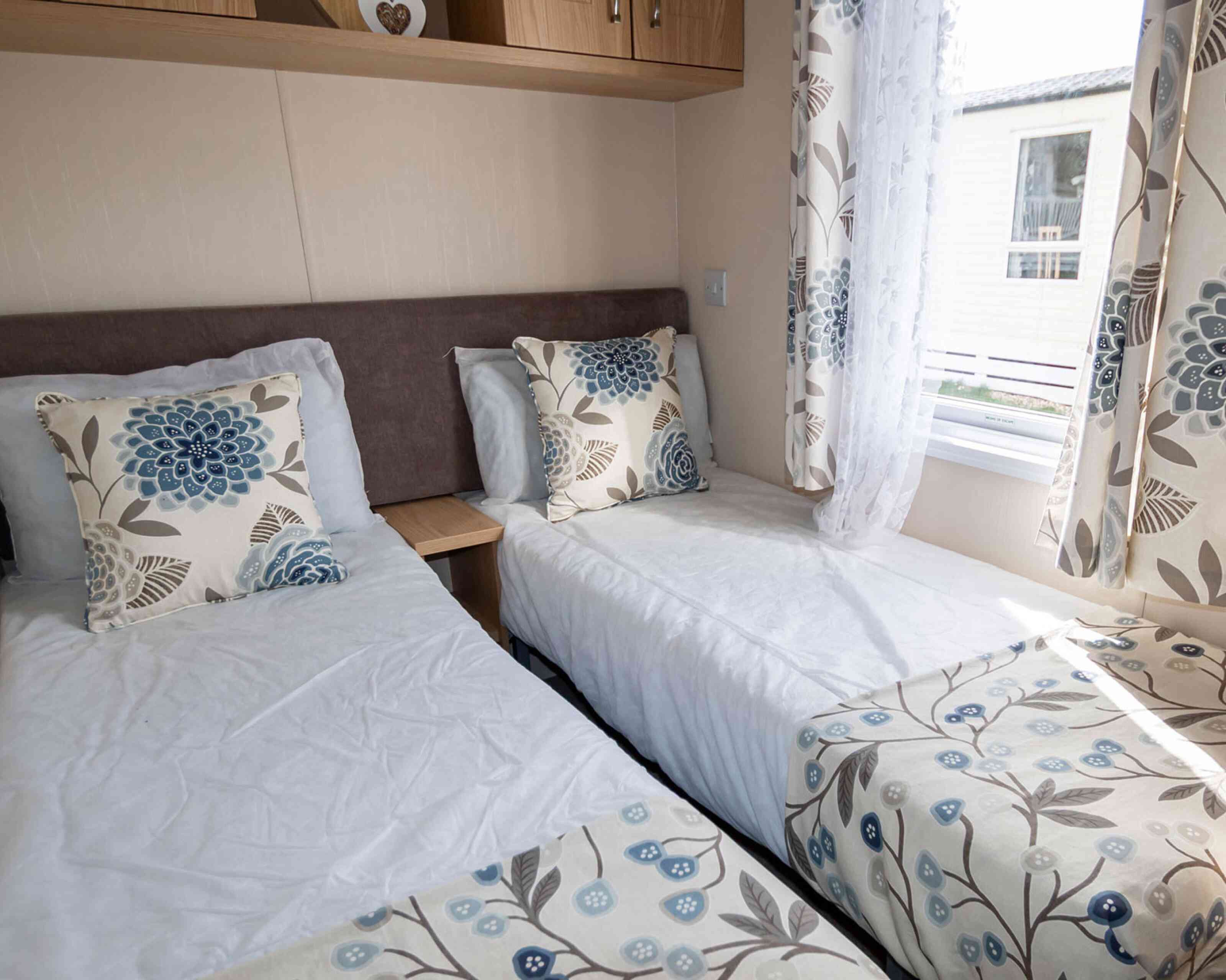 A airy bedroom with two single beds, each adorned with floral cushions and matching comforters. Soft natural light filters through patterned curtains.