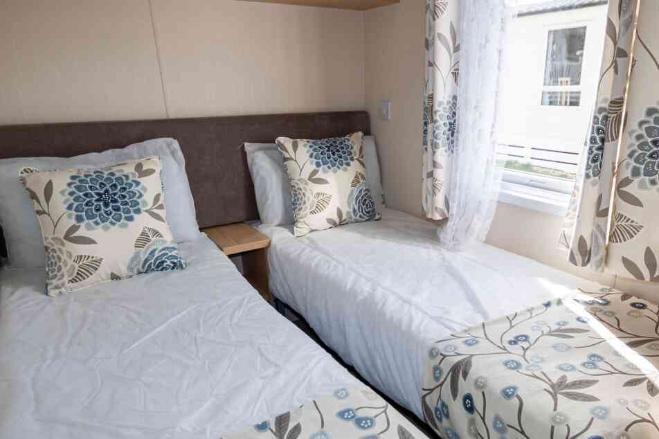 A airy bedroom with two single beds, each adorned with floral cushions and matching comforters. Soft natural light filters through patterned curtains.