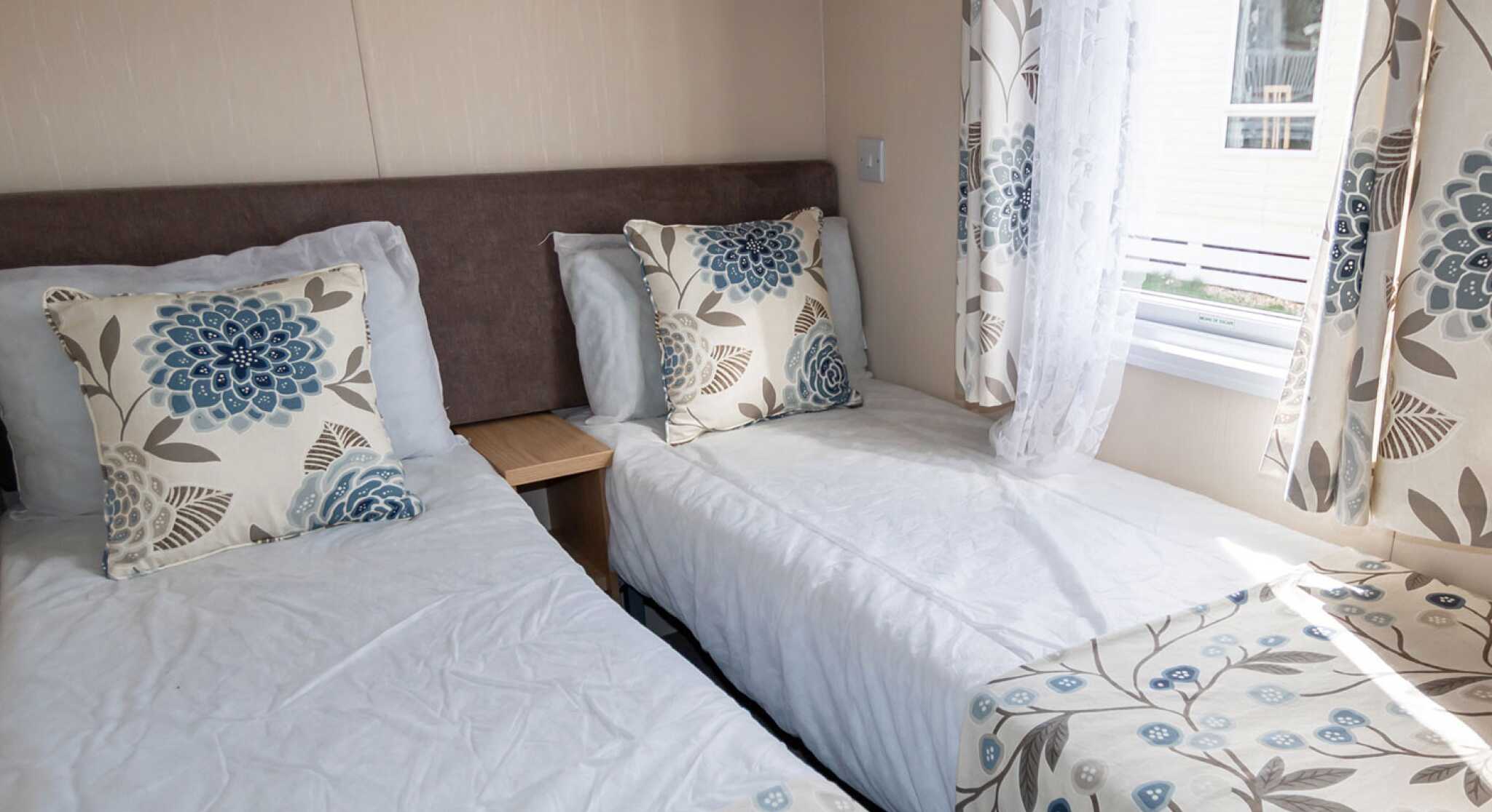 A airy bedroom with two single beds, each adorned with floral cushions and matching comforters. Soft natural light filters through patterned curtains.