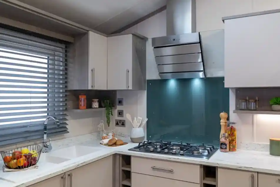 Contemporary kitchen with light cabinetry, a white marble-style countertop, and a teal glass backsplash behind a stainless steel gas cooktop and extractor hood. The kitchen includes a built-in sink below a window with modern horizontal blinds, open shelving with decorative items, and various kitchen essentials like utensils, a fruit basket, and jars